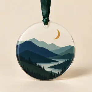Round acrylic ornament printed with navy, teal and green mountains and an ochre crescent moon, on a dark green ribbon