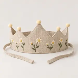 Beige Linen Daisy Crown with Pompom with an embroidered band of small daisies in cream and yellow with green stems on a beige linen crown with a Pompom