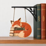 Reading Fox Bookshelf Sign with flat rust-and-cream fox curled around books, hanging by two chains from a thin black wood bracket